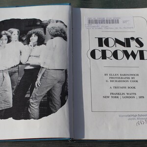 May include: An open book with a black and white photograph on the left page and text on the right. The book's title is "TONI'S CROWD" by Ellen Babinowich, published in 1978. The photo shows a group of people.
