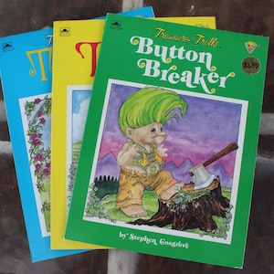 May include: Three vintage children's books with colorful covers. The green book features a troll character with an axe, titled "Button Breaker." The other books have blue and yellow covers. The books are stacked on a dark surface.