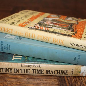 May include: A stack of three vintage books with worn covers. The top book, "Secret of the Old Post Box," has an orange and yellow cover. Below it is a light blue book, and the bottom book is titled "Mutiny in the Time Machine."