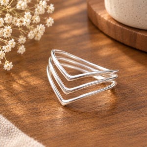 Peut inclure: Une bague en argent présentant un design à trois anneaux avec une forme en chevron pointue. La bague est composée de trois bandes d'argent distinctes, lisses et polies, qui convergent vers un point central, créant un profil en V superposé.