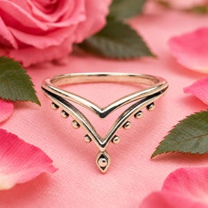 Puede incluir: Un anillo de plata en forma de chevrón con un diseño de doble banda. La banda inferior está adornada con una serie de pequeñas esferas metálicas espaciadas uniformemente, que culminan en un dije central en forma de gota con un punto.