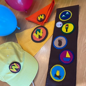 May include: A brown felt sash with colorful embroidered patches, including a campfire, a sailboat, a footprint, a traffic light, and mountains. There are also yellow and orange felt patches with the letter "W" on them. The sash is surrounded by balloons, a yellow felt hat, and a yellow felt patch with the letter "W" on it.