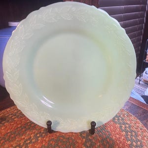 May include: A pale green jadeite dinner plate featuring a scalloped edge and a delicate embossed leaf and berry pattern encircling the rim. The translucent glass plate has a smooth, glossy finish and a circular center with a raised decorative border.