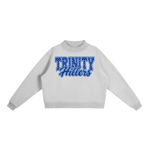 May include: A light gray crewneck sweatshirt featuring the text TRINITY Hillers in blue. The word TRINITY is written in a bold, block-style font with white polka dots, while the word Hillers is written in a blue cursive script font.