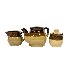 May include: A ceramic tea set consisting of a creamer, a teapot, and a lidded sugar bowl. Each piece features a two-tone design with a dark brown upper section and a light tan lower section, finished with a smooth, glossy glaze throughout.