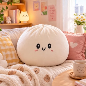 May include: A round, off-white plush pillow shaped like a steamed bun, featuring a simple embroidered face with black oval eyes, a small curved smile, and two small pink blush marks on the cheeks, with a pleated top detail.