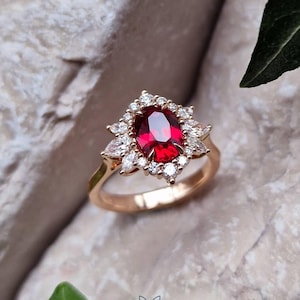 Peut inclure: Une bague en or avec une pierre précieuse rouge foncé de taille ovale au centre, entourée d'un groupe de petites pierres claires de taille marquise et ronde. L'anneau est en or poli et le sertissage forme un motif en étoile autour.