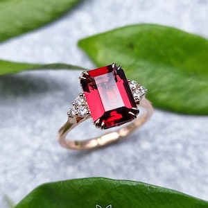 Op de afbeelding: Een roségouden ring met een grote, rechthoekige, smaragdgeslepen dieprode edelsteen in het midden. De ring is aan weerszijden geaccentueerd met een cluster van drie kleine, ronde, helderwitte steentjes die in de ringband zijn gezet.