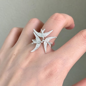 May include: A silver-toned ring featuring three stylized birds arranged in a circular formation. Each bird has detailed wing textures and is positioned with wings outstretched, creating a cohesive, layered design that wraps around the finger.