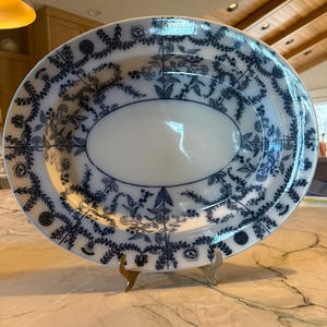 May include: An oval ceramic serving platter featuring a white center and a wide border decorated with a dark blue floral and vine pattern. The platter is displayed on a small gold-toned metal stand, showcasing intricate botanical illustrations.