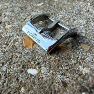 Könnte beinhalten: Eine polierte silberfarbene Uhrenschnalle aus Metall mit dem eingravierten Markennamen FORTIS auf der Vorderseite sowie einem kleinen Kronensymbol. Die Schnalle verfügt über einen geschwungenen Rahmen und einen Dorn zur Befestigung.