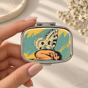 May include: A rectangular metal pill case featuring a graphic illustration of a figure with large butterfly wings curled in a fetal position. The artwork uses a teal background with yellow brushstroke accents and a silver-toned metal frame.