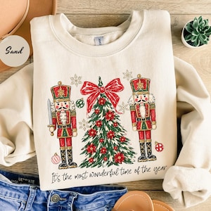 May include: A sand-colored crewneck sweatshirt featuring a graphic of two nutcrackers flanking a decorated Christmas tree with a large red bow. The text "It's the most wonderful time of the year" is printed in black script below the design.