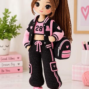 Puede incluir: Una muñeca de ganchillo con cabello castaño en una coleta alta, que viste una chaqueta universitaria negra y rosa, un top corto con el número 28 y la letra A, pantalones cargo negros con ribete rosa y zapatillas rosas y blancas, con mochila negra.
