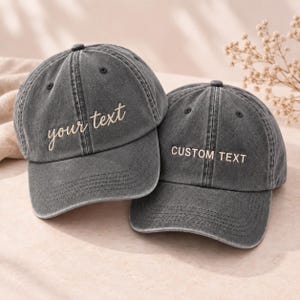 Könnte beinhalten: Zwei anthrazitfarbene Baseballkappen mit verwaschener Optik. Die linke Kappe ist mit dem in Schreibschrift gestickten Text 'your text' in hellem Beige versehen, während die rechte Kappe den in Blockbuchstaben gestickten Text 'CUSTOM TEXT' zeigt.