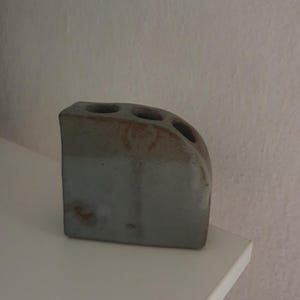 Op de afbeelding: Een kleine, rechthoekige keramische vaas met een gebogen bovenrand en drie ronde openingen. Het object heeft een tweekleurige afwerking, met een lichtgrijs onderste gedeelte en een textuurrijke, bruin-beige glazuurlaag aan de bovenzijde.