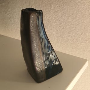 Może przedstawiać: Wysoki, asymetryczny wazon ceramiczny z ciemnobrązowym, teksturowanym matowym wykończeniem z jednej strony oraz błyszczącym, reaktywnym szkliwem w odcieniach głębokiego błękitu, bieli i czerni z drugiej. Wazon posiada wąski, nieregularny otwór u góry.