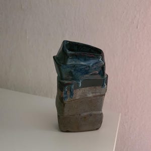 Może przedstawiać: Mały, prostokątny wazon ceramiczny o nieregularnym, ręcznie formowanym kształcie. Górna część pokryta jest błyszczącym, głębokim niebieskim szkliwem, które ścieka na dolną sekcję o matowej, nakrapianej, ziemisto-szarej i brązowej teksturze.