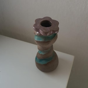 Puede incluir: Un portavelas de cerámica con un cuerpo texturizado en forma de reloj de arena de color marrón, con un acento de esmalte azul verdoso en espiral. La parte superior tiene forma de flor de seis pétalos en tono malva con una abertura central.