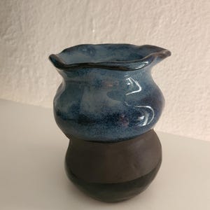 Może przedstawiać: Mały ceramiczny wazon o pękatym kształcie z falistym, rozszerzającym się brzegiem. Górna część pokryta jest błyszczącą, nakrapianą niebieską glazurą, natomiast dolna sekcja wykończona jest matowym, ciemnoszarym kolorem, tworząc dwukolorowy wygląd.