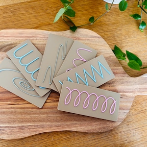 May include: A collection of rectangular tan cards, each featuring a raised, colourful line design. The designs include light blue, pink, and grey squiggles, spirals, and zig-zag patterns. These cards are arranged in a fan shape on a wooden surface.