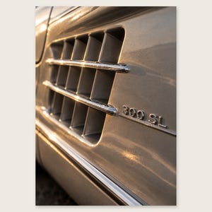 Può includere: Un pannello laterale di un'auto color argento con una presa d'aria rettangolare dotata di tre lamelle cromate orizzontali. La superficie metallica presenta l'emblema cromato 300 SL a destra della presa d'aria, riflettendo la luce solare.