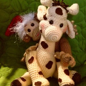 May include: A crocheted giraffe toy with brown spots, sitting on a green surface. A smaller crocheted monkey toy with a red hat is sitting on the giraffe's back. The text "Hooking To Please By Gloria" is visible at the bottom of the image.