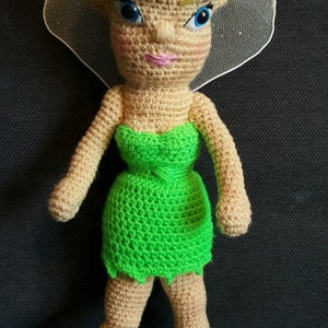 May include: A crocheted Tinkerbell doll with blonde hair, a green dress, and green shoes. The doll is standing on a black surface. "Hooking To Please By Gloria" is written on the surface.