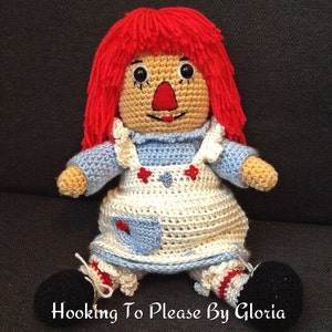 Könnte beinhalten: Eine gehäkelte Stoffpuppe mit roten Wollhaaren, einem blau-weißen Kleid und schwarzen Schuhen. Die Puppe sitzt auf einer dunkelgrauen Oberfläche. Der Text "Hooking To Please By Gloria" ist am unteren Rand des Bildes sichtbar.