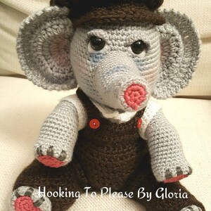May include: A crocheted gray elephant wearing a brown cap and brown overalls with red buttons. The elephant has pink feet and a pink nose. The text "Hooking To Please By Gloria" is visible in the image.