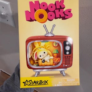 May include: A yellow product box for Nook Nooks by Starbox featuring a graphic of a red vintage-style television set. Inside the screen, a white rabbit figure sits in a yellow armchair with a remote, a lamp, a clock, and a bowl of carrots.
