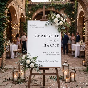 May include: A white wedding welcome sign on a wooden easel featuring the text Welcome to the wedding of Charlotte and Harper, Ashford Manor, September 14th 20XX, and We're so glad you're here, decorated with white flowers and green foliage.