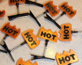 May include: A collection of hair clips featuring orange flame-shaped toppers with the word HOT printed in black. Each topper is attached to a black metal alligator clip by a flexible metal spring, allowing the flame design to bounce and move.