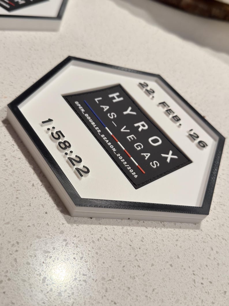 May include: A hexagonal white and black plaque featuring the text HYROX LAS VEGAS, 22, FEB, 26, and 1:58:22. The centre includes a black trapezoid with the text OPEN_DOUBLES_SEASON_2025/2026 and a small horizontal blue, white, and red line graphic.