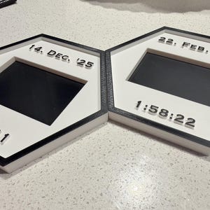 May include: Two hexagonal frames with black borders and white centres. The left frame displays 14. DEC. '25 and 2:13:11. The right frame displays 22. FEB. '26 and 1:58:22. Each frame features a central rectangular black cutout for a photo or display.