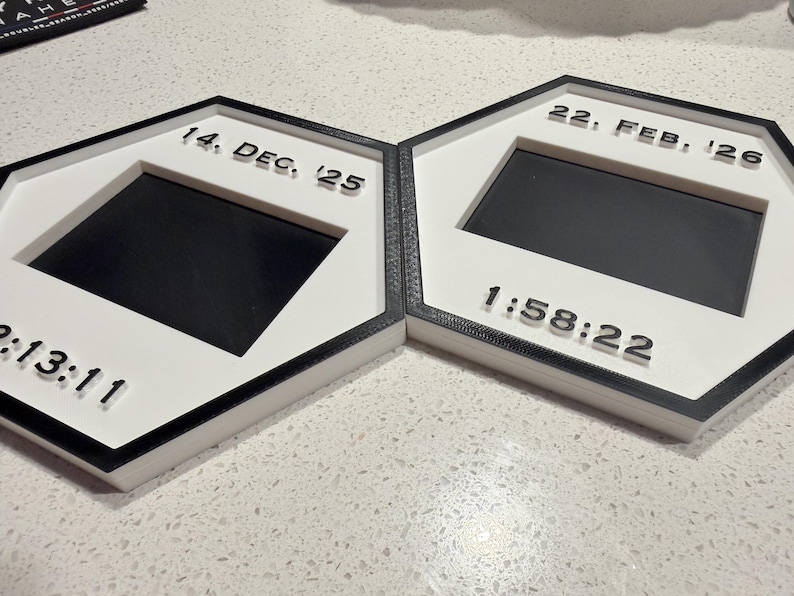 May include: Two hexagonal frames with black borders and white centres. The left frame displays 14. DEC. '25 and 2:13:11. The right frame displays 22. FEB. '26 and 1:58:22. Each frame features a central rectangular black cutout for a photo or display.