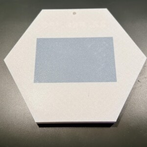 May include: A white hexagonal ornament featuring a centred light blue rectangular patch. The ornament has a small circular hole at the top edge for hanging. The surface of the white material appears matte and smooth, while the blue rectangle is solid.