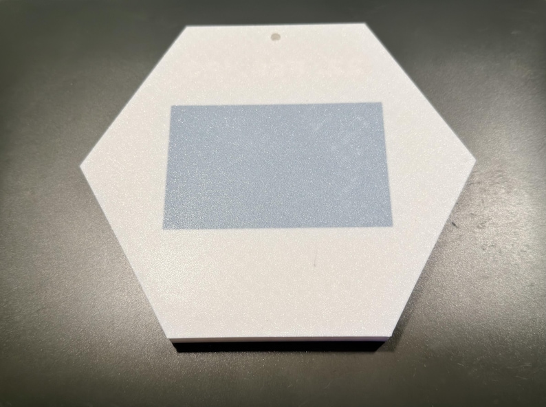 May include: A white hexagonal ornament featuring a centred light blue rectangular patch. The ornament has a small circular hole at the top edge for hanging. The surface of the white material appears matte and smooth, while the blue rectangle is solid.