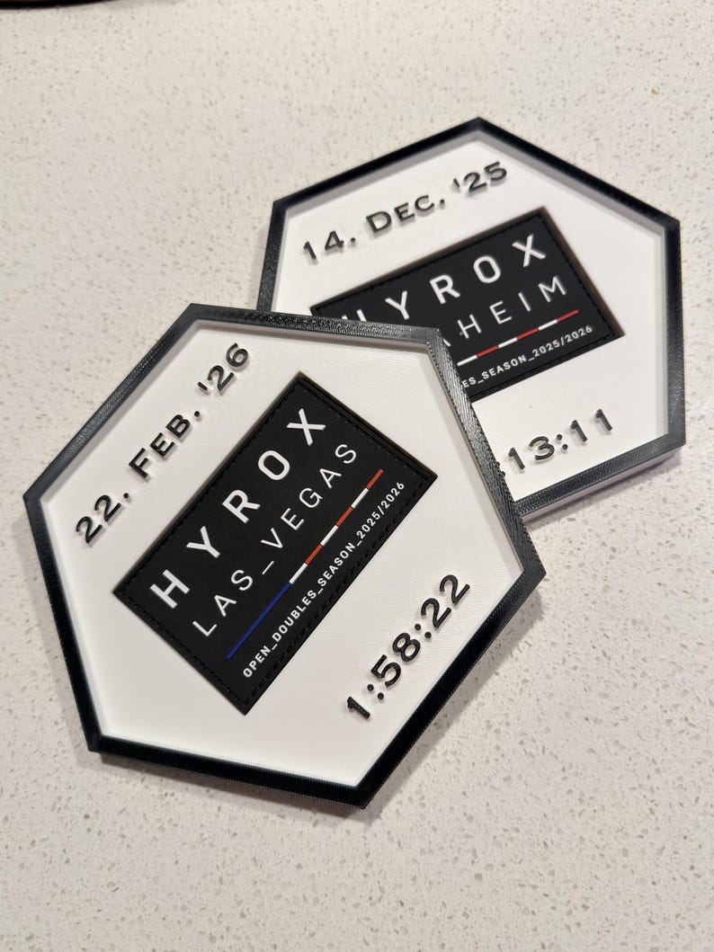 May include: Two hexagonal white plaques with black borders featuring raised black text. One reads 22. FEB. '26, HYROX LAS_VEGAS, OPEN_DOUBLES_SEASON_2025/2026, and 1:58:22. The other reads 14. DEC. '25, HYROX MANNHEIM, ES_SEASON_2025/2026, and 13:11.