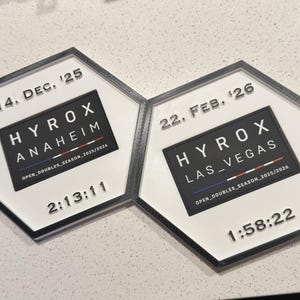 May include: Two hexagonal plaques with black frames and white centres. The left plaque reads 14. DEC. '25, HYROX ANAHEIM, OPEN_DOUBLES_SEASON_2025/2026, and 2:13:11. The right plaque reads 22. FEB. '26, HYROX LAS_VEGAS, OPEN_DOUBLES_SEASON_2025/2026, and 1:58:22.