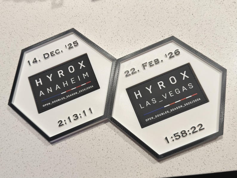 May include: Two hexagonal plaques with black frames and white centres. The left plaque reads 14. DEC. '25, HYROX ANAHEIM, OPEN_DOUBLES_SEASON_2025/2026, and 2:13:11. The right plaque reads 22. FEB. '26, HYROX LAS_VEGAS, OPEN_DOUBLES_SEASON_2025/2026, and 1:58:22.