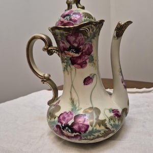 May include: A porcelain teapot featuring a cream-colored base decorated with hand-painted purple poppy flowers and green foliage. The teapot includes a matching lid with a decorative finial, a curved handle, and a tall spout, all accented with gold trim.