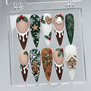 May include: A set of ten press-on nails in a clear plastic case, featuring festive holiday designs including gingerbread figures, white snowflakes, holly berries, pine branches, a bell, and a gingerbread house, with brown and white drip accents.