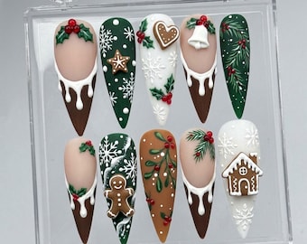 May include: A set of ten press-on nails in a clear plastic case, featuring festive holiday designs including gingerbread figures, white snowflakes, holly berries, pine branches, a bell, and a gingerbread house, with brown and white drip accents.