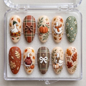 May include: A set of ten almond-shaped press-on nails in a clear plastic case. The designs feature autumn themes including white ducks wearing scarves and sweaters, orange pumpkins, brown plaid patterns with 3D bows, and various fall leaves and flowers.
