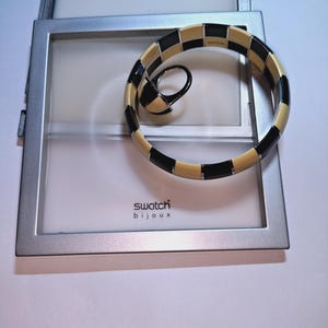 Puede incluir: Un brazalete rígido y un anillo a juego de Swatch Bijoux con un patrón de cuadros repetitivo de segmentos rectangulares alternos en negro y crema, separados por finas divisiones de metal plateado, presentados sobre un marco de exhibición cuadrado.