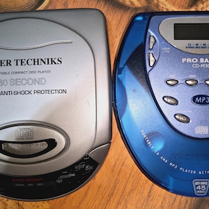 May include: A silver Laser Techniks portable compact disc player with 60 second digital anti-shock protection and a blue Pro Basic CD-M30 portable CD and MP3 player with 45 second anti-shock protection, both featuring control buttons.