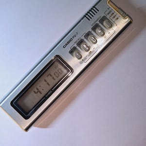 Könnte beinhalten: Ein silberner Casio PQ-7 Reisewecker mit einem rechteckigen Digitaldisplay, das 4:17 PM anzeigt. Das Gerät verfügt über eine metallische Oberfläche, ein Lautsprechergitter und fünf Bedientasten für Stoppuhr-, Alarm- und Timerfunktionen.
