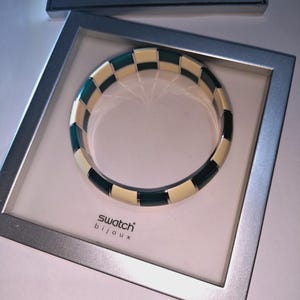 Puede incluir: Una pulsera rígida circular con un patrón alterno de segmentos rectangulares de color crema y verde azulado oscuro. La pulsera se presenta dentro de una caja cuadrada de tono plateado con el texto swatch bijoux impreso en negro sobre la base.