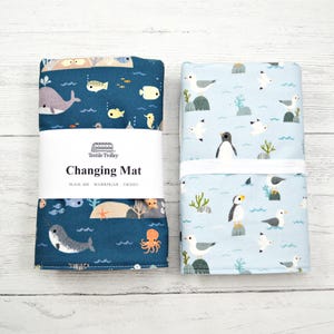 May include: Two rolled baby changing mats. One is navy blue with sea creatures, including a whale, fish, and octopus. The other is light blue with seagulls and penguins. The mats are labeled "Changing Mat" with "Textile Trolley" branding.